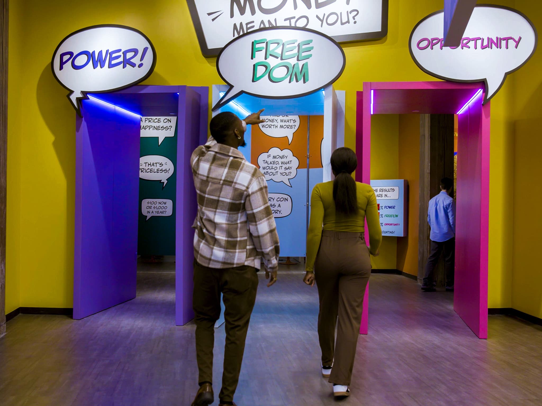 Couple walks through MoMoney's bright corridor past a comic-style speech-bubble exhibit asking what money means.