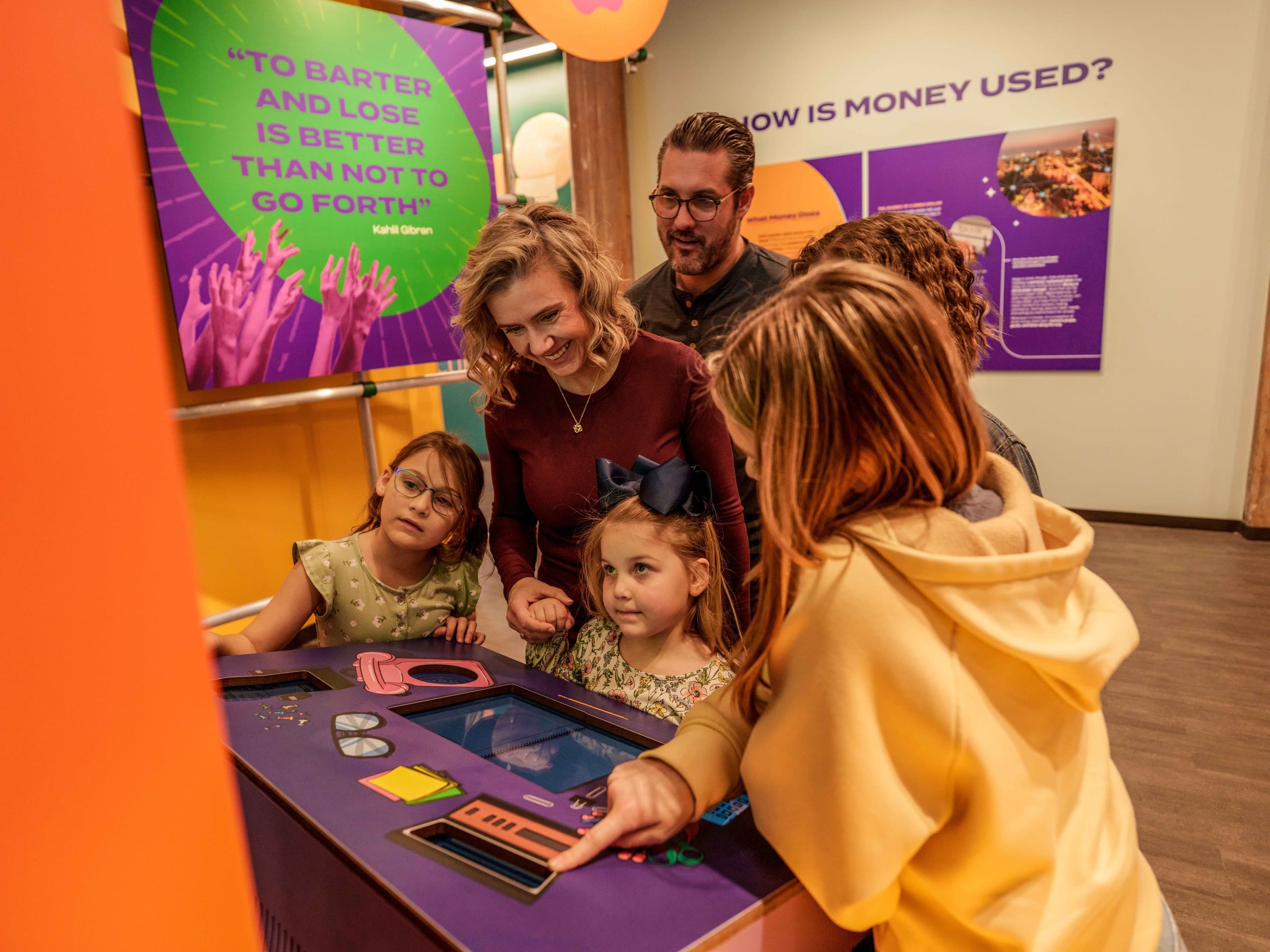 Family with children gathered around MoMoney's interactive barter exhibit, smiling and pointing as they play to win.