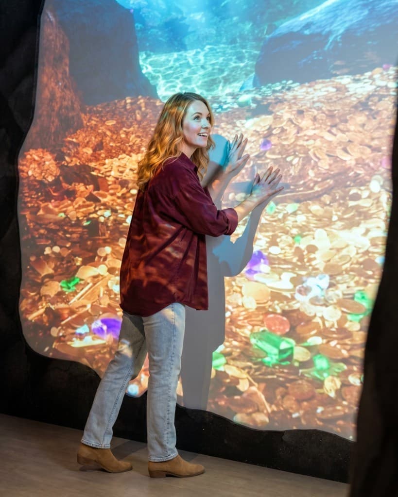 Woman presses her hands against MoMoney's glowing wall of gold coins and gems at the Hoard of Gold exhibit, smiling in wonder.